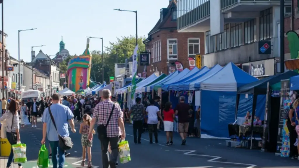 A busy outdoor community street festival on a sunny day, with crowds of people browsing market stalls beneath blue and white gazebos, a colourful bouncy castle visible in the background along a town high street.
