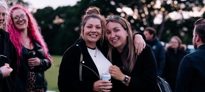 Two smiling women pose together at an outdoor festival, one with her arm around the other. A woman with bright pink hair laughs in the background alongside other festival-goers, with trees visible behind them