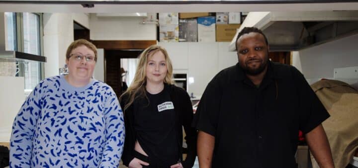 Three people standing together indoors in a community kitchen. The person in the centre is wearing a black top with an Elthorne Pride N19 badge. The woman to the left is wearing a white and blue top and there is a man on the right wearing a black t-shirt