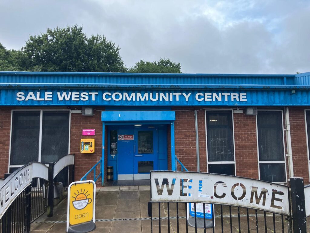 The entrance of Sale West Community Centre, it has a blue entrance door, welcome signs, a defibrillator, and a Sunshine Community Café open sign out front.