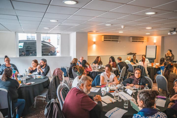 Participants sit in small groups around tables during a workshop, with notebooks, drinks and discussion materials spread out in front of them.