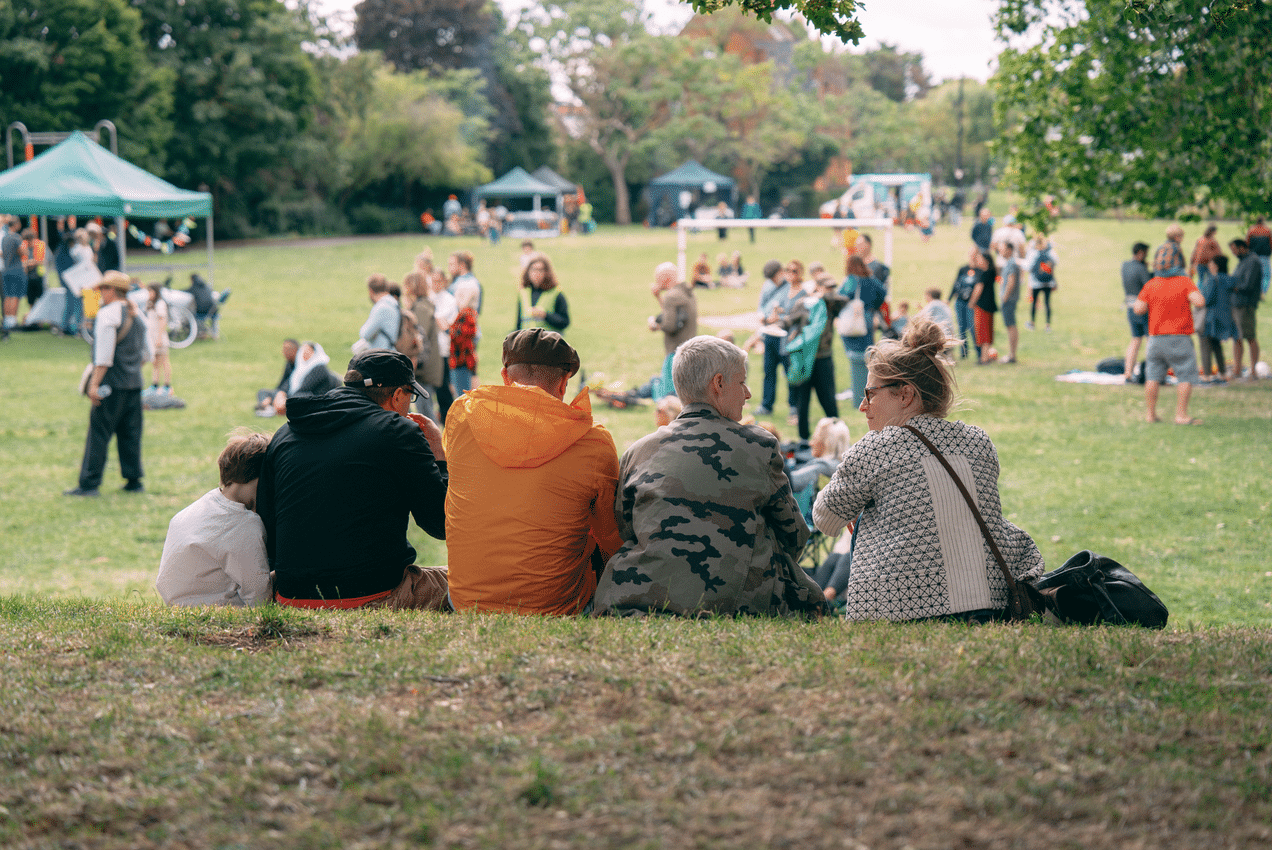 A group of five people sit on the grass with their backs to the camera at an outdoor community event in a park. Around them, small groups of people, gazebos and activities are scattered across the large green field, with trees surrounding the area on a bright day.