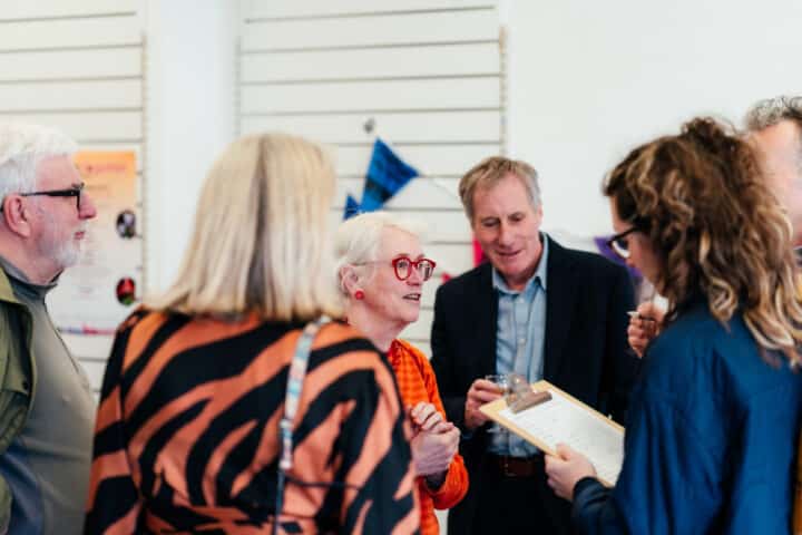 A small group of adults stand talking together indoors at a community event or meeting. In the centre, an older woman with white hair, red glasses and an orange top is speaking, while another person holds a clipboard and takes notes; colourful bunting and posters are visible in the background.