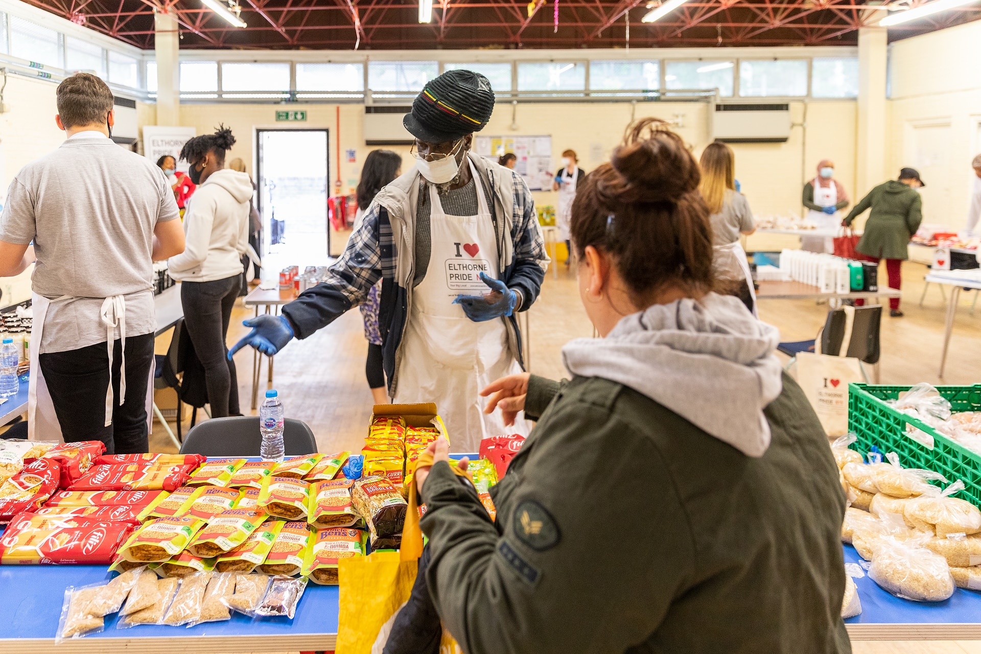 Volunteers and community members gather in a hall during a food distribution event. In the foreground, a volunteer wearing an apron, face mask and gloves gestures towards a table stocked with packaged foods. Other volunteers are visible in the room