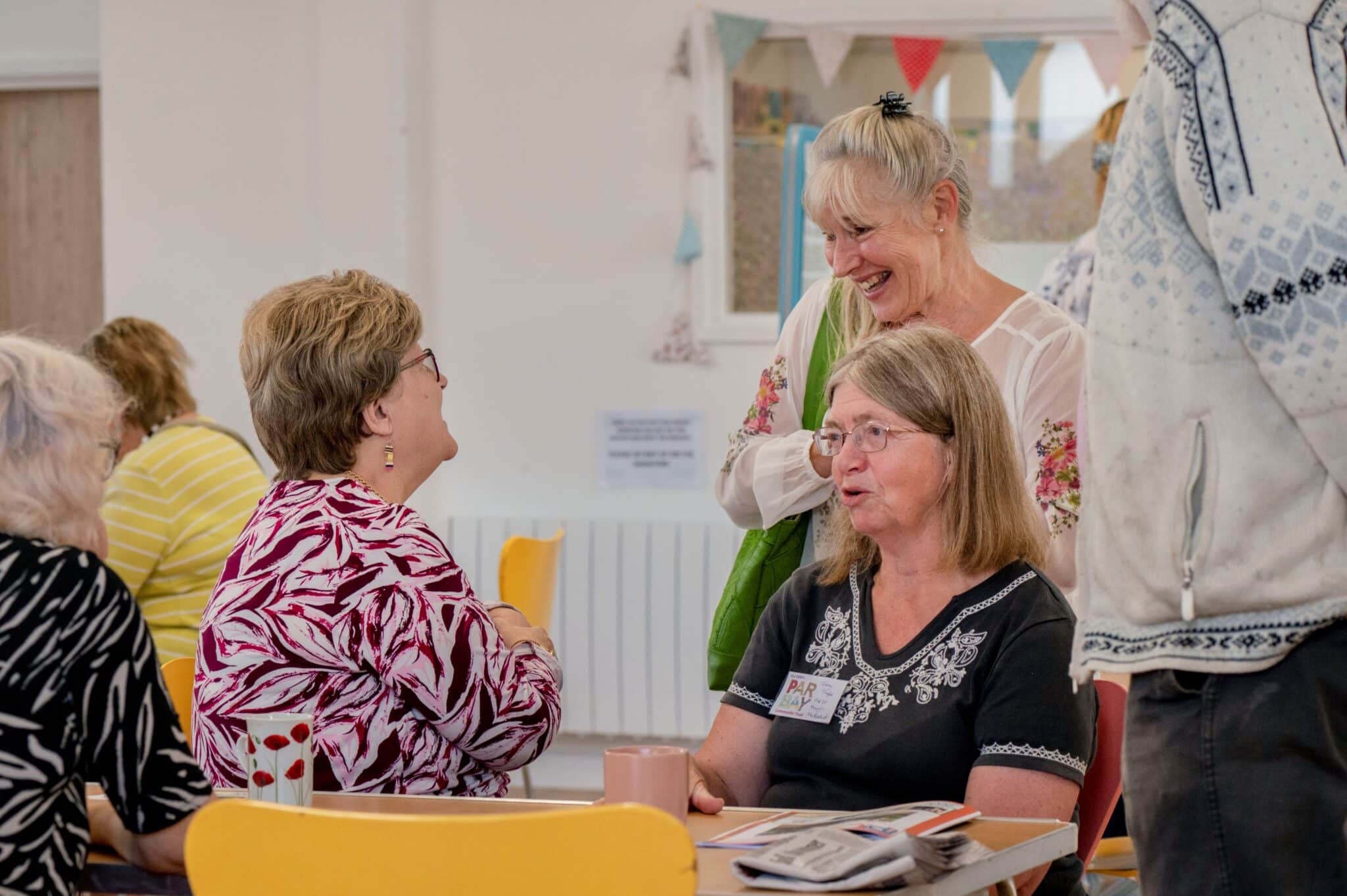 A group of women chat warmly around a table inside the Par Bay community space. A seated woman wearing a PAR Bay name badge speaks with others gathered around her, including a woman in a floral top who is laughing, and a woman in a bold patterned top who faces her across the table.
