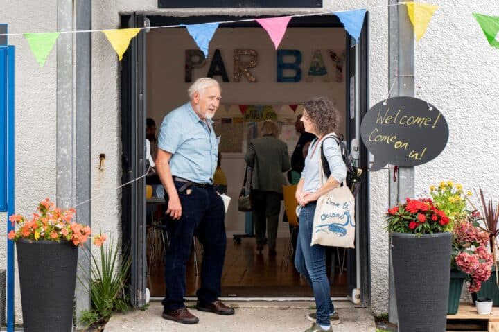 Two people chat in the doorway of the Par Bay community space, decorated with colourful bunting and a chalkboard sign reading 