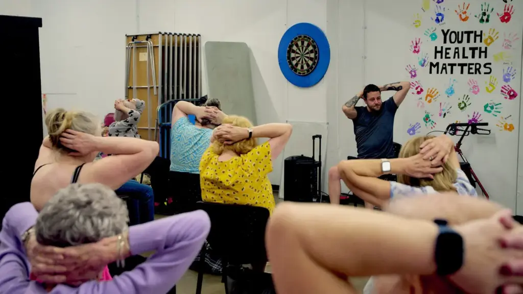 A group of adults sits on chairs in a community hall, following a seated exercise led by an instructor at the front of the room. Participants have their hands behind their heads as they perform a stretching exercise. A colourful wall display reading "Youth Health Matters," decorated with painted handprints, a dartboard, and folded tables in the background suggest a shared community space focused on health and wellbeing.