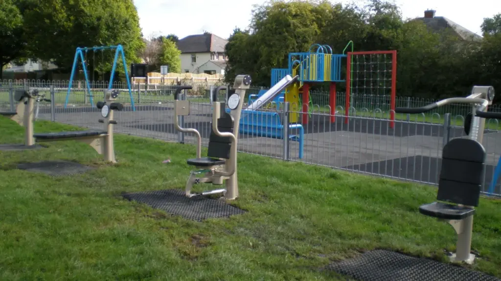 Outdoor gym equipment on grass in the foreground, with a fenced children's playground behind featuring blue swings, a slide, climbing frame, and red climbing net. Trees and houses are visible in the background
