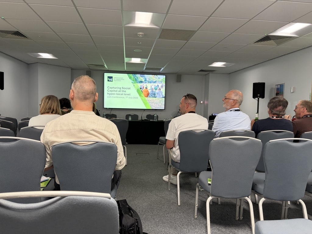 Attendees seated in rows of grey chairs, viewed from behind, watch a presentation on a large screen at the front of a conference room. The screen displays a slide titled 'Capturing Social Capital at the Hyper-Local Level, LGA Conference, 7th July 2026,' alongside a photo of people gathered outdoors in a park