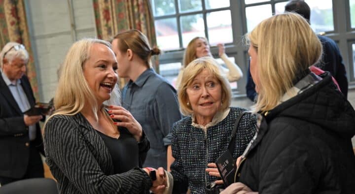 hree people are engaged in conversation at a community event inside a hall. In the foreground, a smiling woman shakes hands with another attendee while an older woman stands beside them, listening attentively. Other people chat in the background near large windows, creating a warm and welcoming atmosphere that reflects community connection and networking.