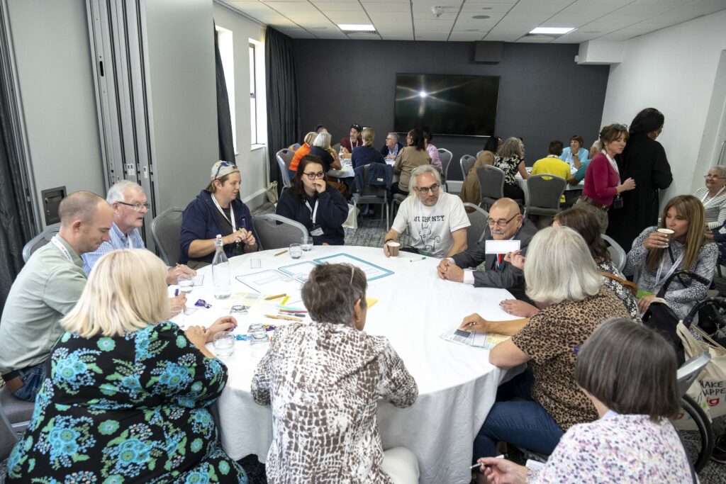 A group of attendees sit around a round table with a white cloth, discussing worksheets and diagrams during a workshop session, with pens, water glasses, and papers spread across the table. Several more round tables of participants are visible in the background of the conference room, with a large screen mounted on the far wall.
