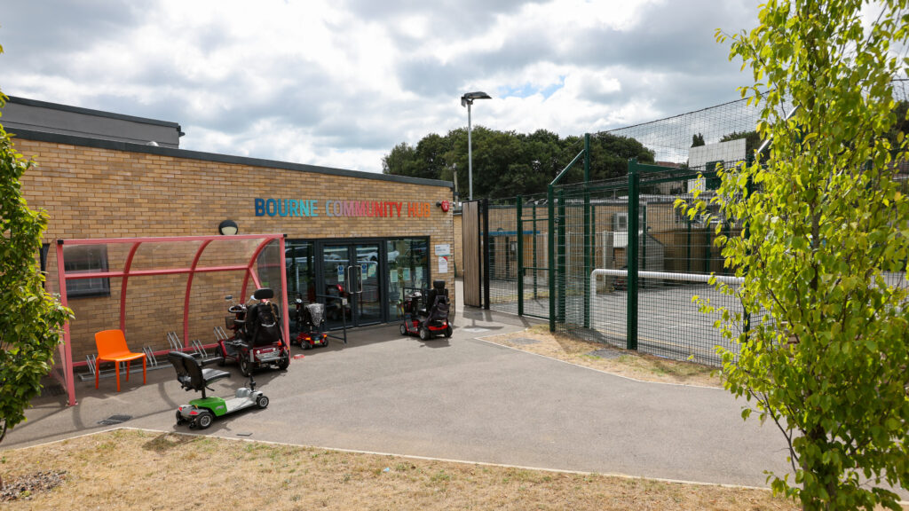 Exterior of Bourne Community Hub, a single-storey brick building with a colourful sign above the entrance. Several mobility scooters are parked outside near a covered bike shelter and an orange chair, with a fenced sports court and trees visible in the background.